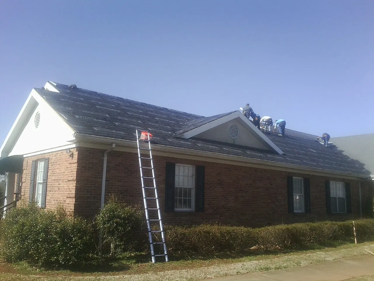 Crew tearing shingles off a brick ranch roof with a ladder set — Emergency Roof Repair work by our crew in State Farm