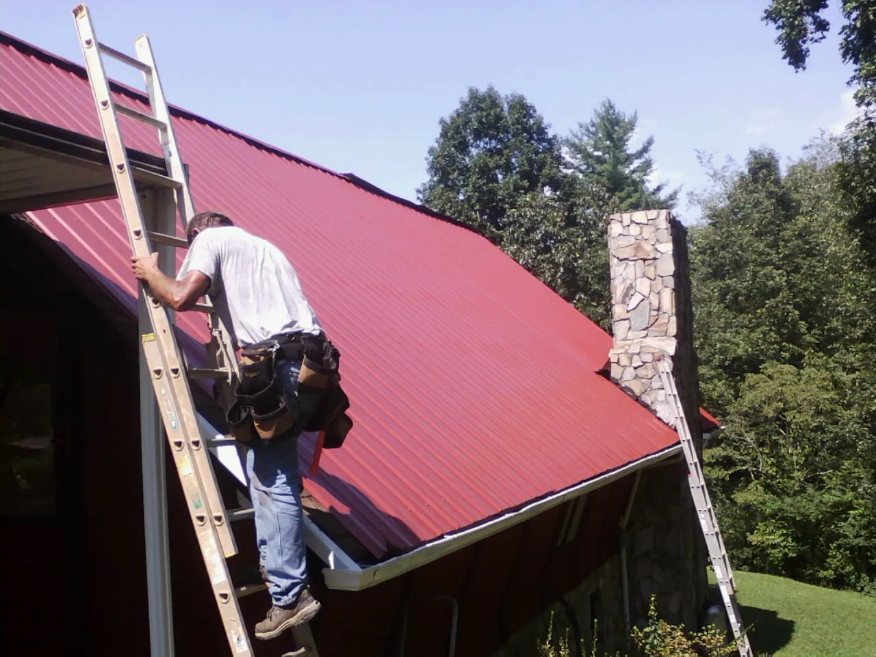 Roofer on a ladder beside a red metal roof and stone chimney — Soffit & Fascia Repair by Roofing Brothers in State Farm